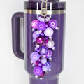 A plum Stanley with a silver clasp and a beaded straw charm. The charm features a cluster of purple beads in various shades and sizes, including a star-shaped bead. The mug has a handle and a dark purple lid.