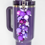 A plum Stanley with a silver clasp and a beaded straw charm. The charm features a cluster of purple beads in various shades and sizes, including a star-shaped bead. The mug has a handle and a dark purple lid.