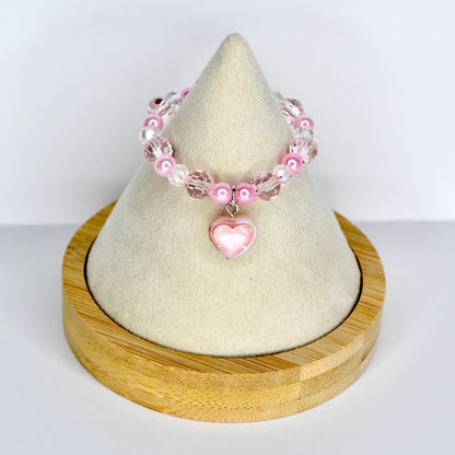 A pink beaded monster doll necklace with a heart charm. The necklace features a mix of pink and clear beads, with a heart-shaped pendant at the center. The necklace is displayed on a cream-colored surface.