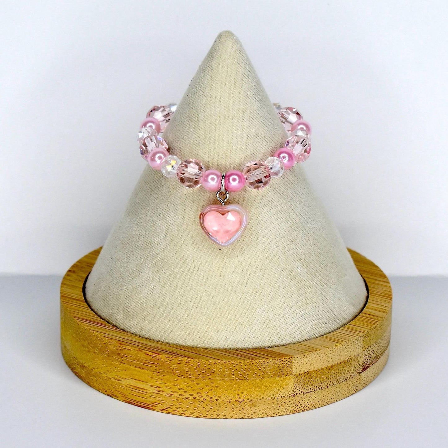 A pink beaded monster doll necklace with a heart charm. The mini necklace features a mix of clear, pink, and pearl-like beads. A pink heart-shaped charm hangs from the center. The necklace is displayed on a cream-colored cone-shaped stand, set on a wooden base.