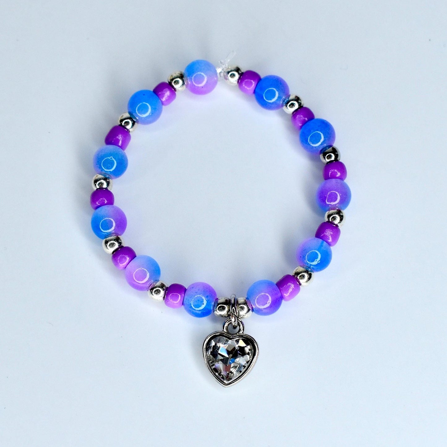 A beaded monster doll necklace featuring blue, purple, and silver beads. The necklace has a heart-shaped charm with a clear, faceted design. The beads are strung on a flexible cord, creating a piece of jewelry.
