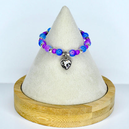 A beaded monster doll necklace with blue, purple, and silver beads. The necklace has a silver heart charm. The necklace is displayed on a white cone-shaped stand with a wooden base.