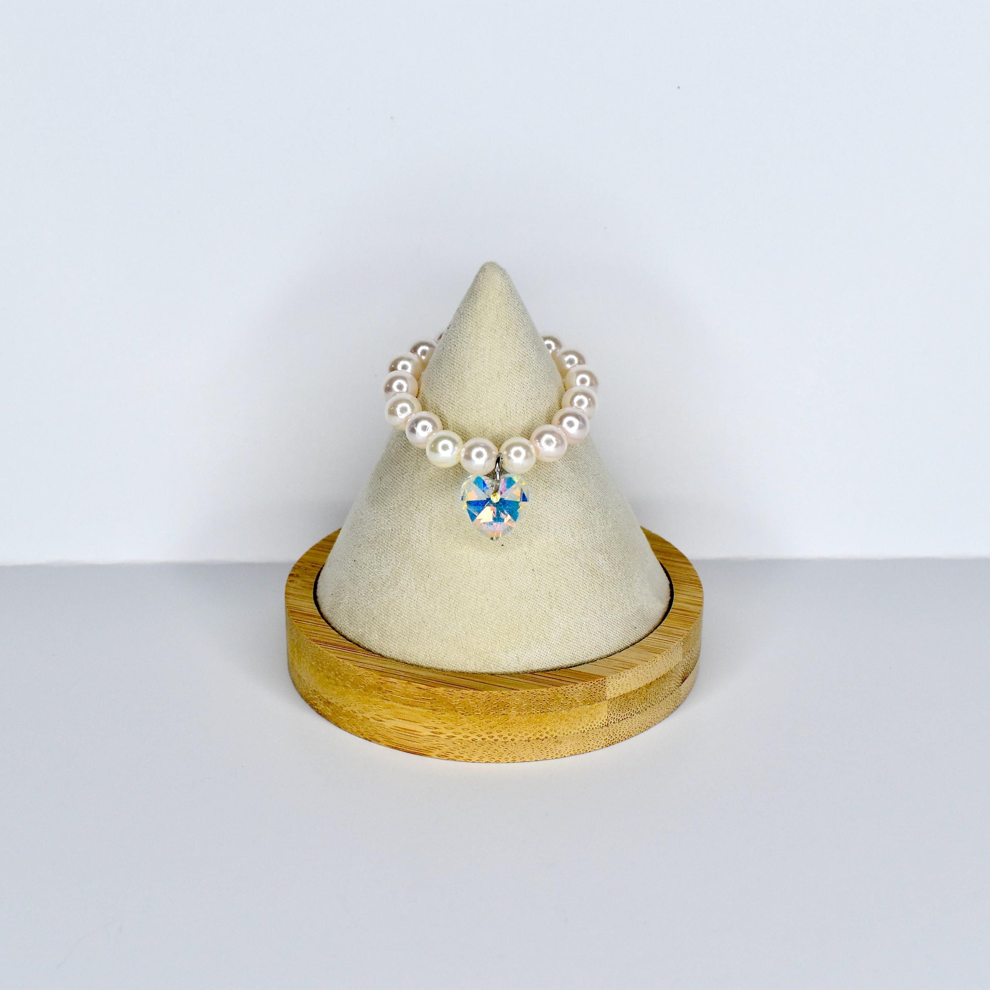 A pearl beaded monster doll necklace with a sparkling, multi-colored crystal pendant. The necklace is displayed on a small, wooden, cone-shaped stand with a cream-colored fabric top. The necklace is a delicate piece of jewelry.
