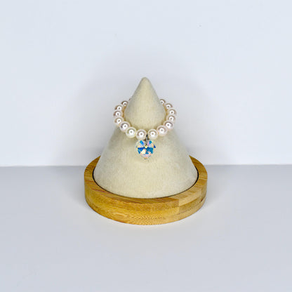 A pearl beaded monster doll necklace with a sparkling, multi-colored crystal pendant. The necklace is displayed on a small, wooden, cone-shaped stand with a cream-colored fabric top. The necklace is a delicate piece of jewelry.