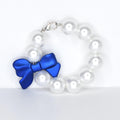 A pearl tumbler handle bracelet with a vibrant blue bow. The bracelet is composed of large, round, off-white pearls. The bow is made of a smooth, satin-like material and is positioned on the bracelet.