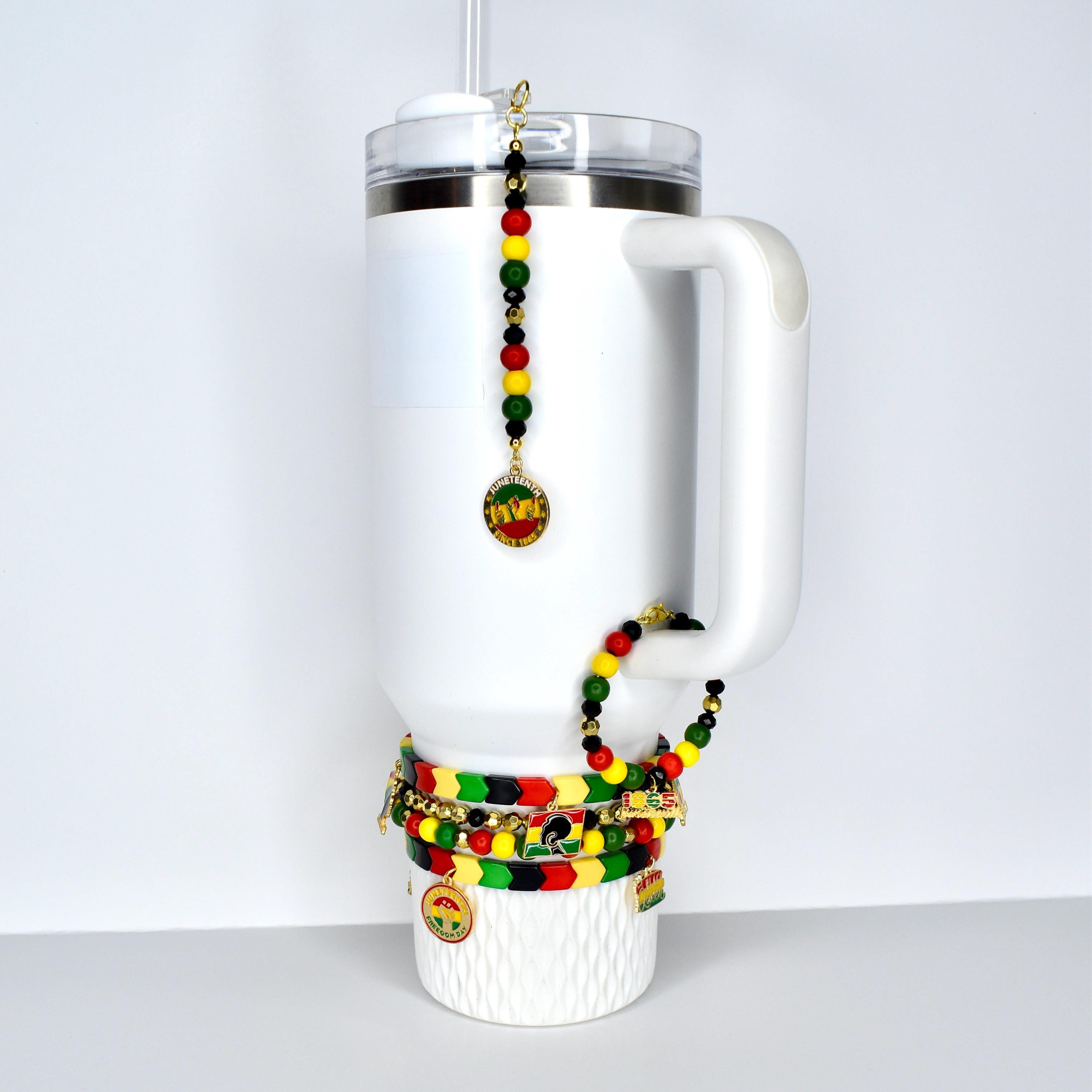 White insulated tumbler with a handle, adorned with beaded bracelets and a charm. The bracelets feature red, yellow, green, and black beads. A charm with a gold-colored pendant hangs from the lid. The tumbler has a straw.