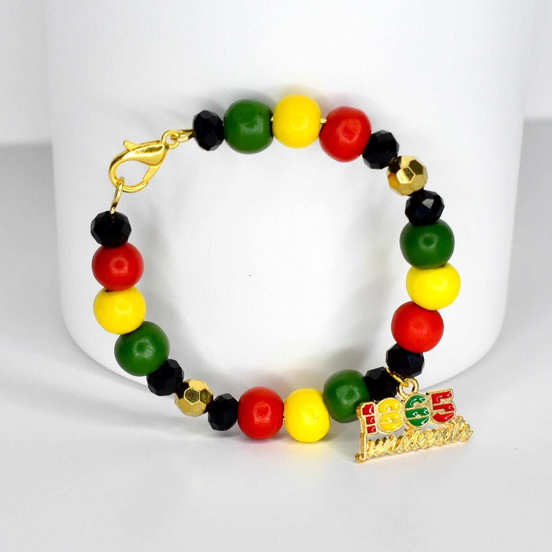 A beaded bracelet featuring red, yellow, green, and black beads. The bracelet includes a gold-colored charm with the text "1865 Juneteenth". The bracelet is on a white background.