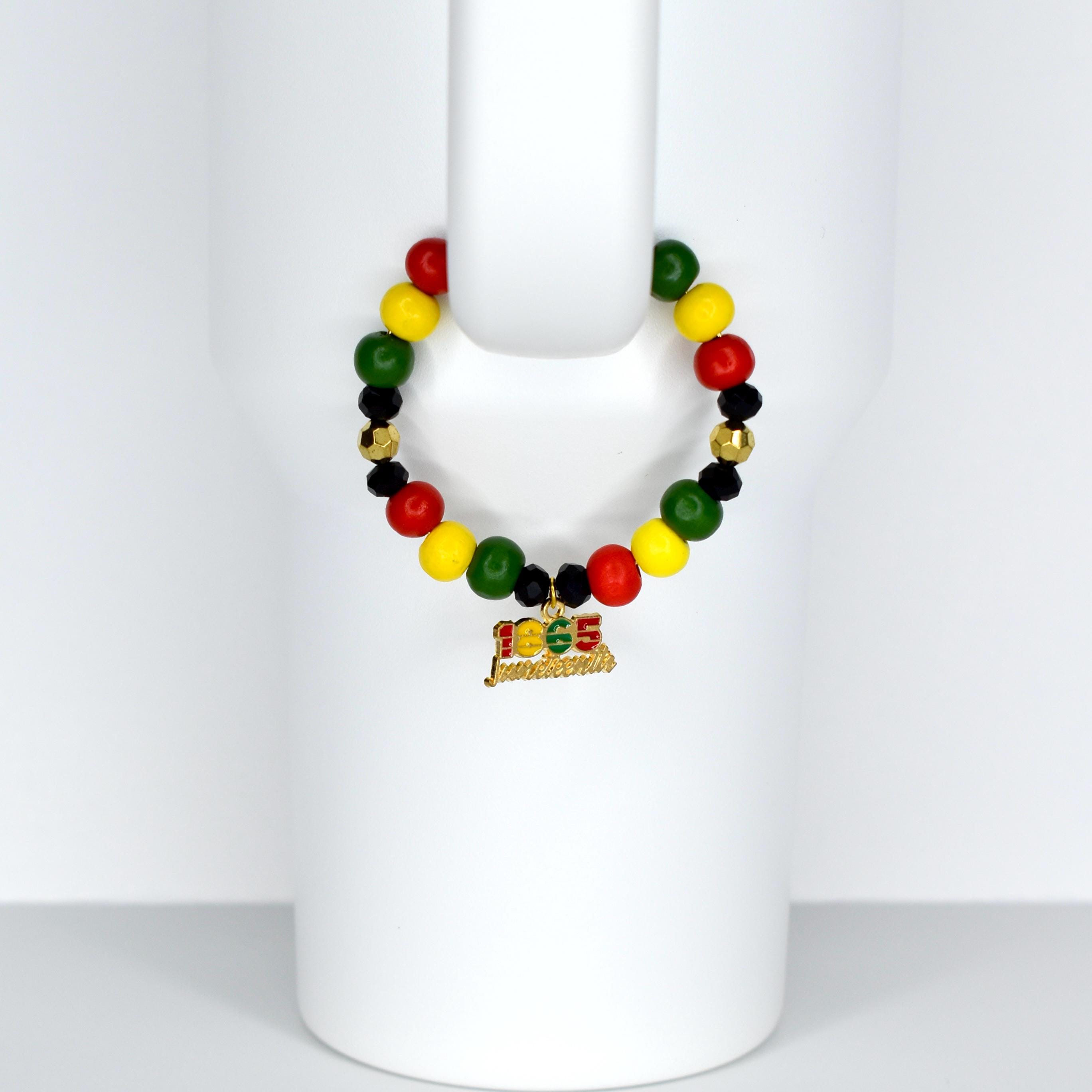 A beaded tumbler handle bracelet featuring red, yellow, green, black, and gold beads. A gold charm with the text "1865 Juneteenth" hangs from the bracelet. The bracelet is displayed on a white surface.