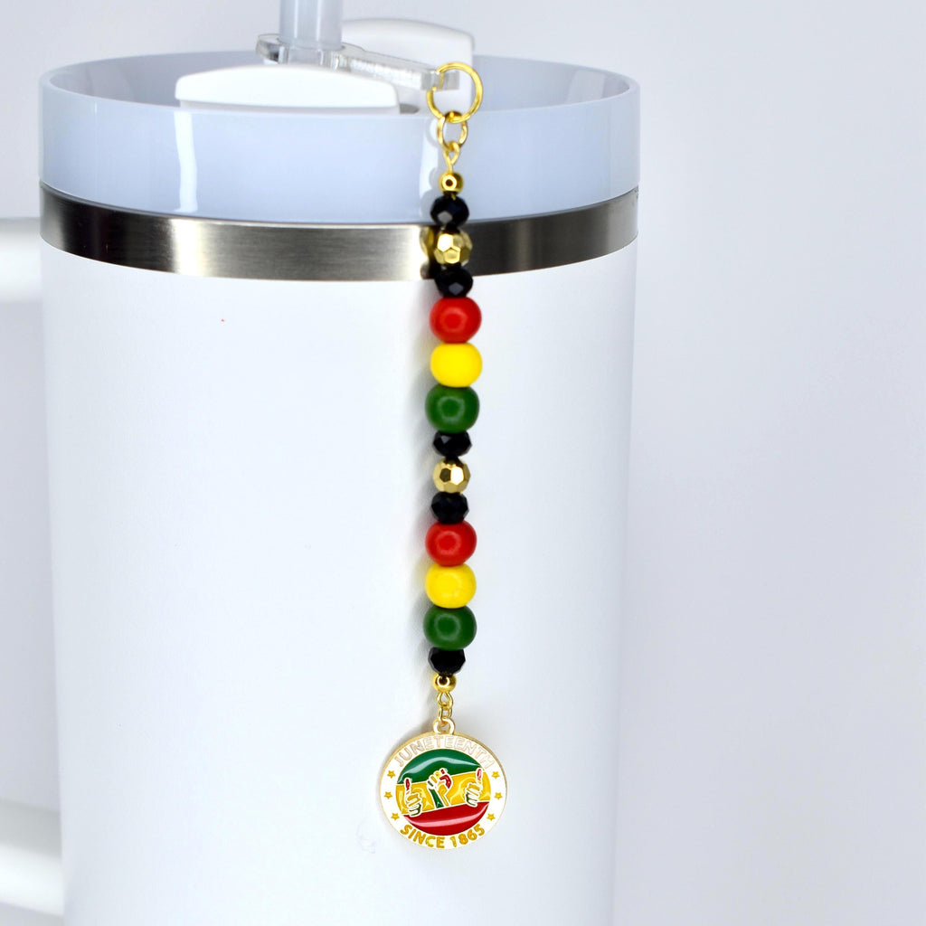 A white tumbler with a Juneteenth straw charm. The charm features a series of colorful beads in red, yellow, green, and black, and a circular pendant with text and a graphic. The text on the pendant reads "SINCE 1865".