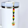 A white tumbler with a Juneteenth straw charm. The charm features a series of colorful beads in red, yellow, green, and black, and a circular pendant with text and a graphic. The text on the pendant reads "SINCE 1865".
