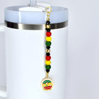 A white tumbler with a Juneteenth straw charm. The charm features a series of colorful beads in red, yellow, green, and black, and a circular pendant with text and a graphic. The text on the pendant reads "SINCE 1865".