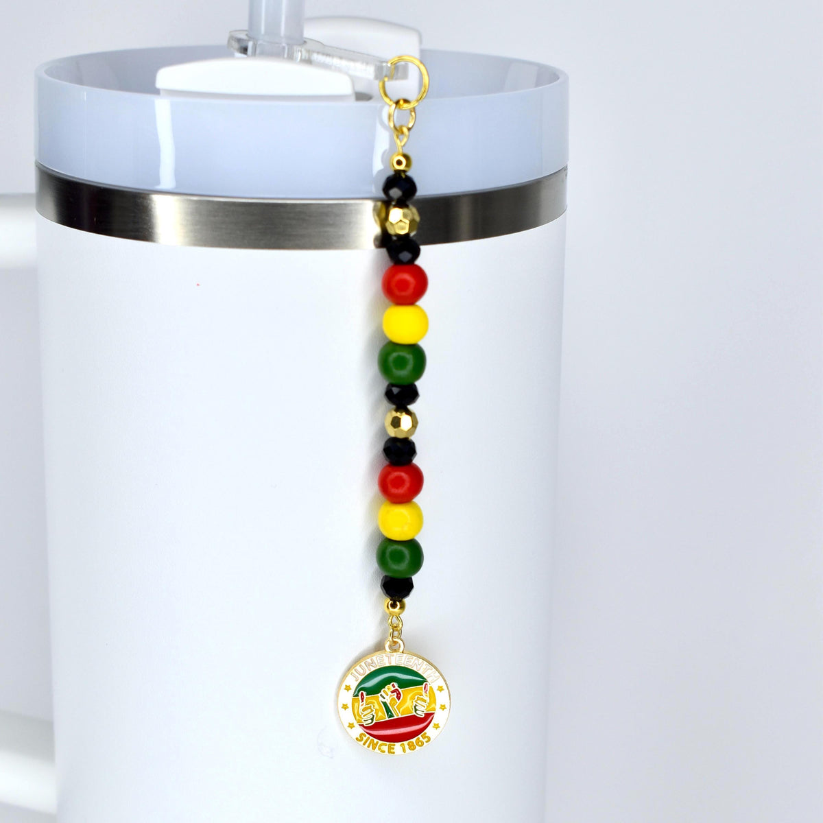 A white tumbler with a Juneteenth straw charm. The charm features a series of colorful beads in red, yellow, green, and black, and a circular pendant with text and a graphic. The text on the pendant reads "SINCE 1865".