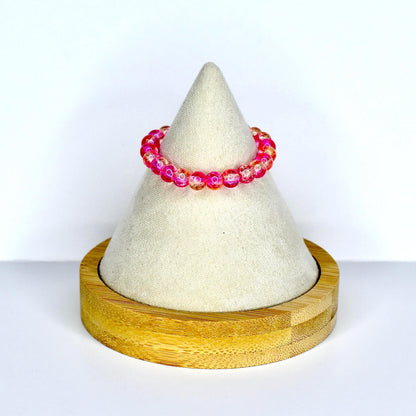 A vibrant pink beaded monster doll necklace with a mix of clear and orange beads. The necklace is displayed on a white cone-shaped stand, which is set on a round wooden base. The necklace is a stylish accessory.