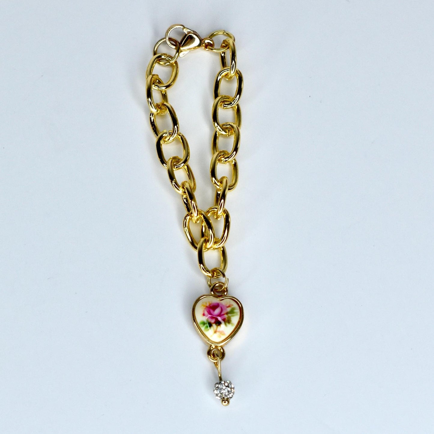A gold-colored chain monster doll necklace with a heart-shaped charm. The charm features a floral design with pink and green accents. A small, sparkling crystal dangles below the heart. The necklace is on a white background.