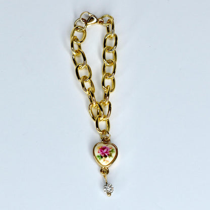A gold-colored chain monster doll necklace with a heart-shaped charm. The charm features a floral design with pink and green accents. A small, sparkling crystal dangles below the heart. The necklace is on a white background.