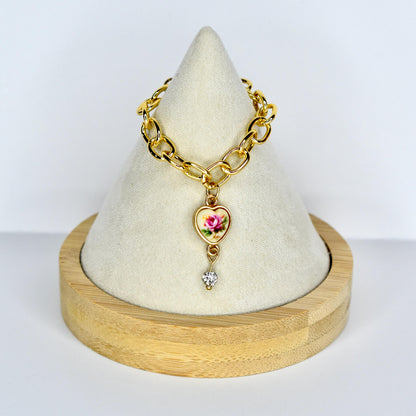 Gold-toned chain monster doll necklace with a heart-shaped pendant. The pendant features a pink rose design on a cream background, with a small, clear gemstone dangling below. The mini necklace is displayed on a white, cone-shaped stand.