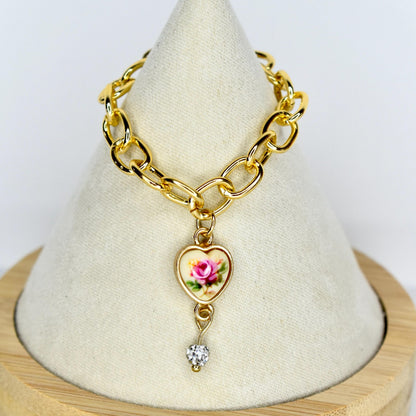 Gold-toned chain 17cm monster doll necklace with a heart-shaped pendant. The pendant features a pink rose design on a cream background, framed by a gold border. A small, clear, heart-shaped gem hangs below the pendant. The necklace is displayed on a white surface.