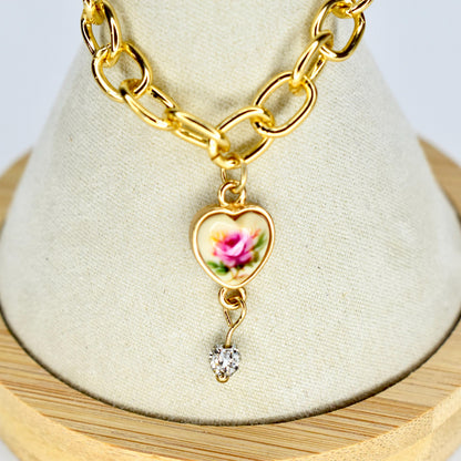 Gold-toned chain necklace with a heart-shaped pendant. The pendant features a pink rose design on a cream background, with a small, sparkling heart charm dangling below. The chain has large, oval links.