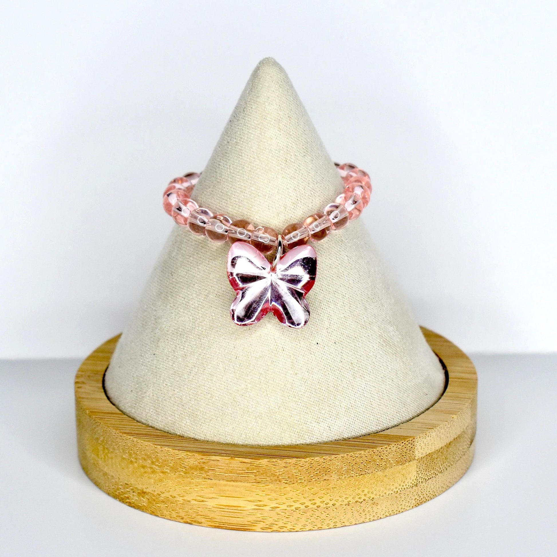 A light pink beaded monster doll necklace with a butterfly charm. The necklace features small, round, translucent pink beads. The butterfly charm is a solid pink color with a metallic sheen. The necklace is displayed on a white cone-shaped stand.