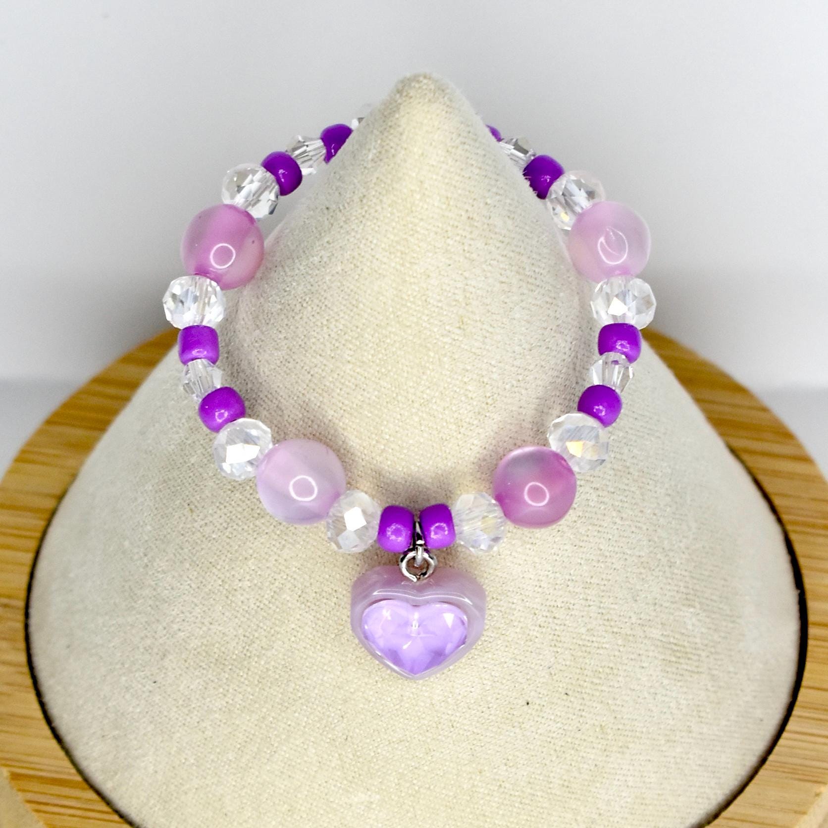 A beaded mini necklace featuring a heart pendant. The necklace is composed of purple, clear, and pink beads. The heart pendant is a light purple color. The necklace is displayed on a cream-colored surface.