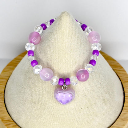A beaded mini necklace featuring a heart pendant. The necklace is composed of purple, clear, and pink beads. The heart pendant is a light purple color. The necklace is displayed on a cream-colored surface.