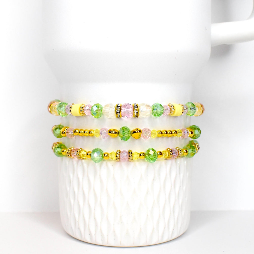 Three beaded bracelets with yellow, green, pink, and clear beads. The bracelets are accented with gold-colored beads and spacers. The bracelets are displayed on a white ceramic mug.
