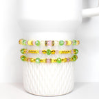 Three beaded bracelets with yellow, green, pink, and clear beads. The bracelets are accented with gold-colored beads and spacers. The bracelets are displayed on a white ceramic mug.