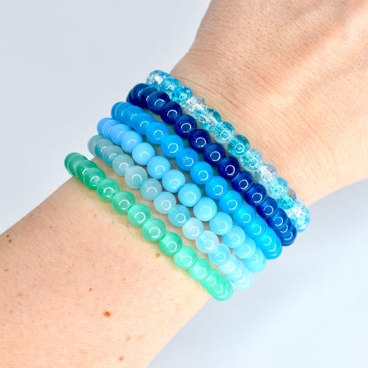 A collection of six beaded bracelets in shades of blue and green. The bracelets are stacked on a wrist, showcasing a gradient of colors from light green to deep blue. The beads are round and vary in size.