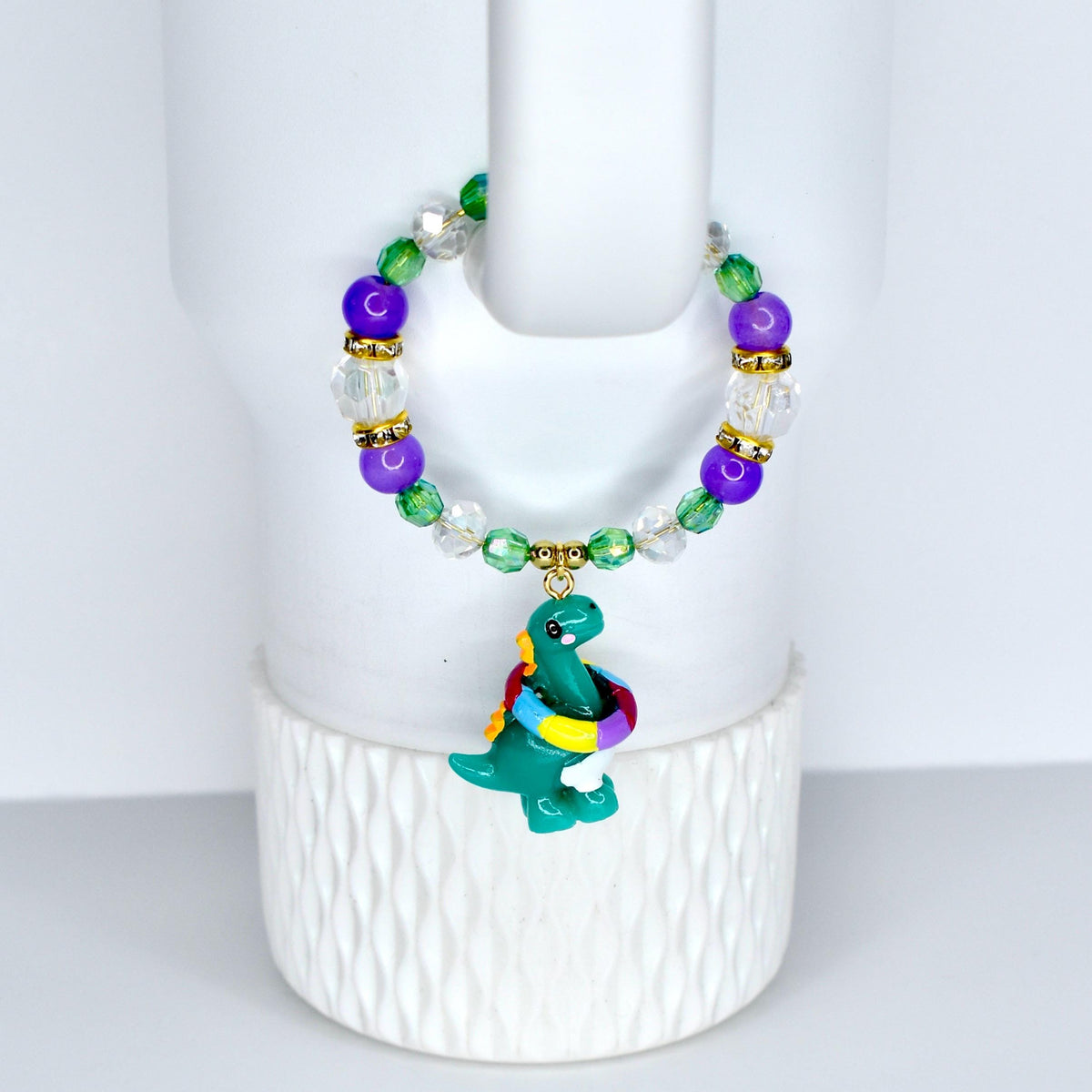 A beaded bracelet with a green dinosaur charm. The bracelet features purple, green, and clear beads with gold accents.