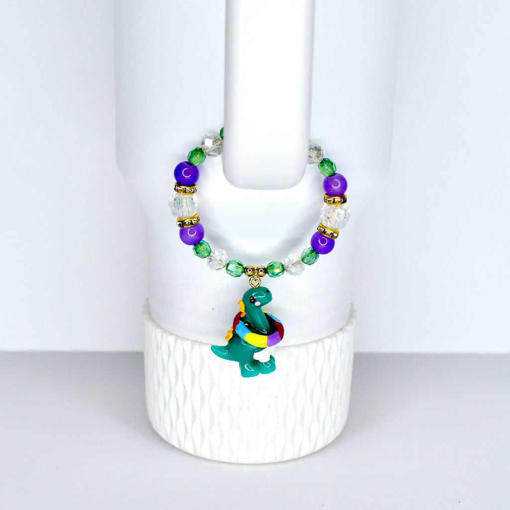 A beaded bracelet featuring a green dinosaur charm with a colorful scarf. The bracelet has purple, green, clear, and gold-colored beads. The dinosaur charm is approximately 1 inch in height.