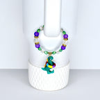 A beaded bracelet featuring a green dinosaur charm with a colorful scarf. The bracelet has purple, green, clear, and gold-colored beads. The dinosaur charm is approximately 1 inch in height.