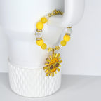 A yellow beaded tumbler handle charm with a gold clasp and a smiling sun charm. The bracelet features yellow and clear beads, with a gold sun pendant wearing sunglasses. The charm is approximately 1 inch in diameter.