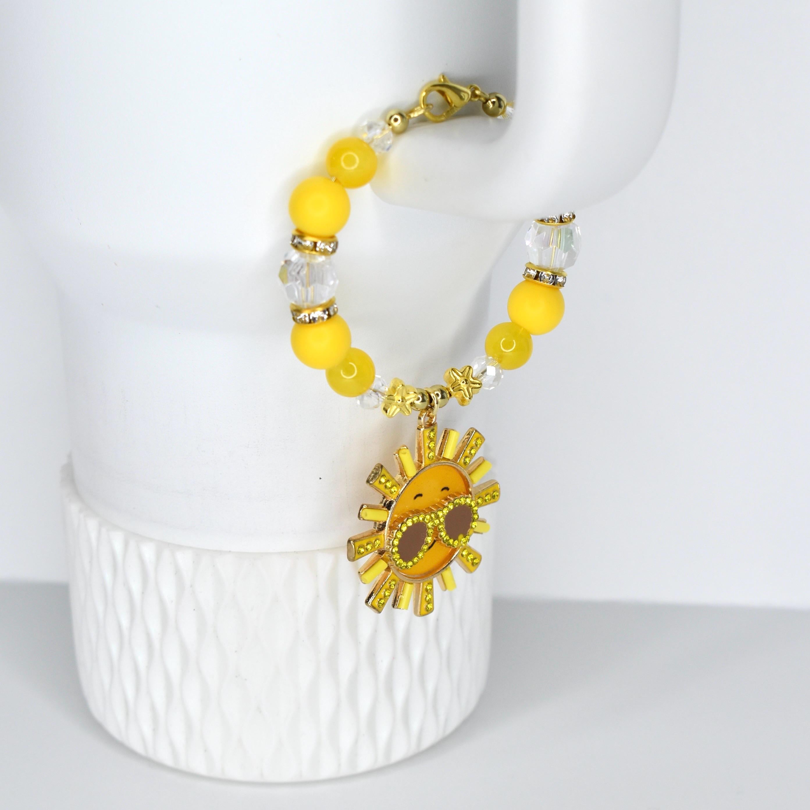 A yellow beaded tumbler handle charm with a gold clasp and a smiling sun charm. The bracelet features yellow and clear beads, with a gold sun pendant wearing sunglasses. The charm is approximately 1 inch in diameter.