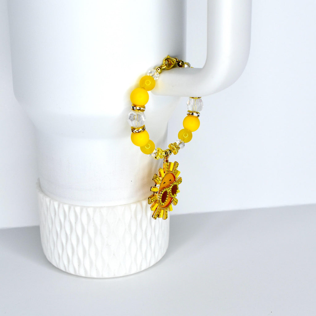 A yellow beaded tumbler handle bracelet with a gold sun charm. The bracelet features yellow and clear beads, with a gold clasp. The sun charm has a smiling face and is surrounded by gold rays.
