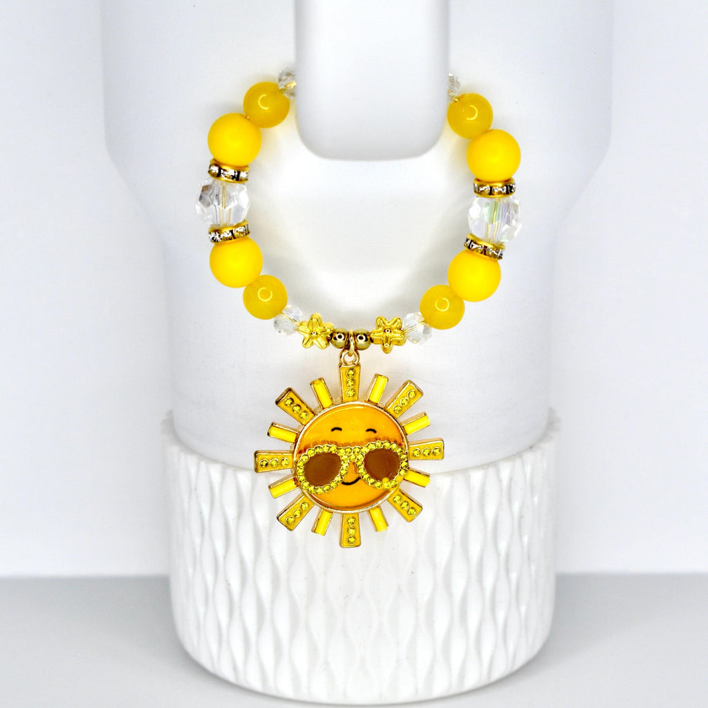 A yellow beaded tumbler handle bracelet with a gold sun charm. The charm features a smiling sun wearing sunglasses. The handle bracelet has yellow and clear beads with gold accents. The bracelet is displayed on a white, textured surface.