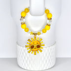 A yellow beaded tumbler handle bracelet with a gold sun charm. The charm features a smiling sun wearing sunglasses. The handle bracelet has yellow and clear beads with gold accents. The bracelet is displayed on a white, textured surface.