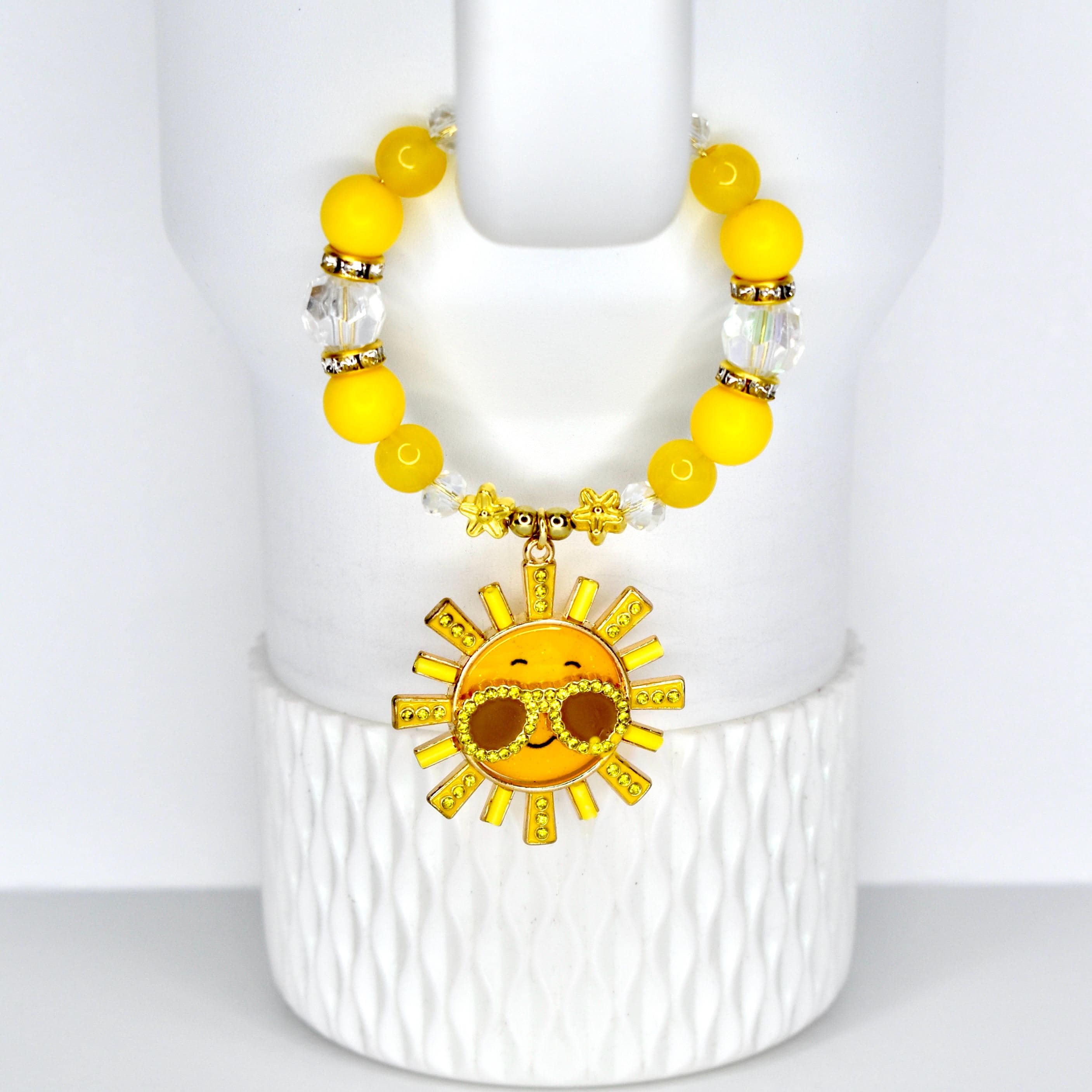 A yellow beaded tumbler handle bracelet with a gold sun charm. The charm features a smiling sun wearing sunglasses. The handle bracelet has yellow and clear beads with gold accents. The bracelet is displayed on a white, textured surface.