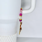 A gold-colored charm with pink and purple gemstones and a beaded strand of pink, white, and purple beads. The charm is hanging on a white mug handle.