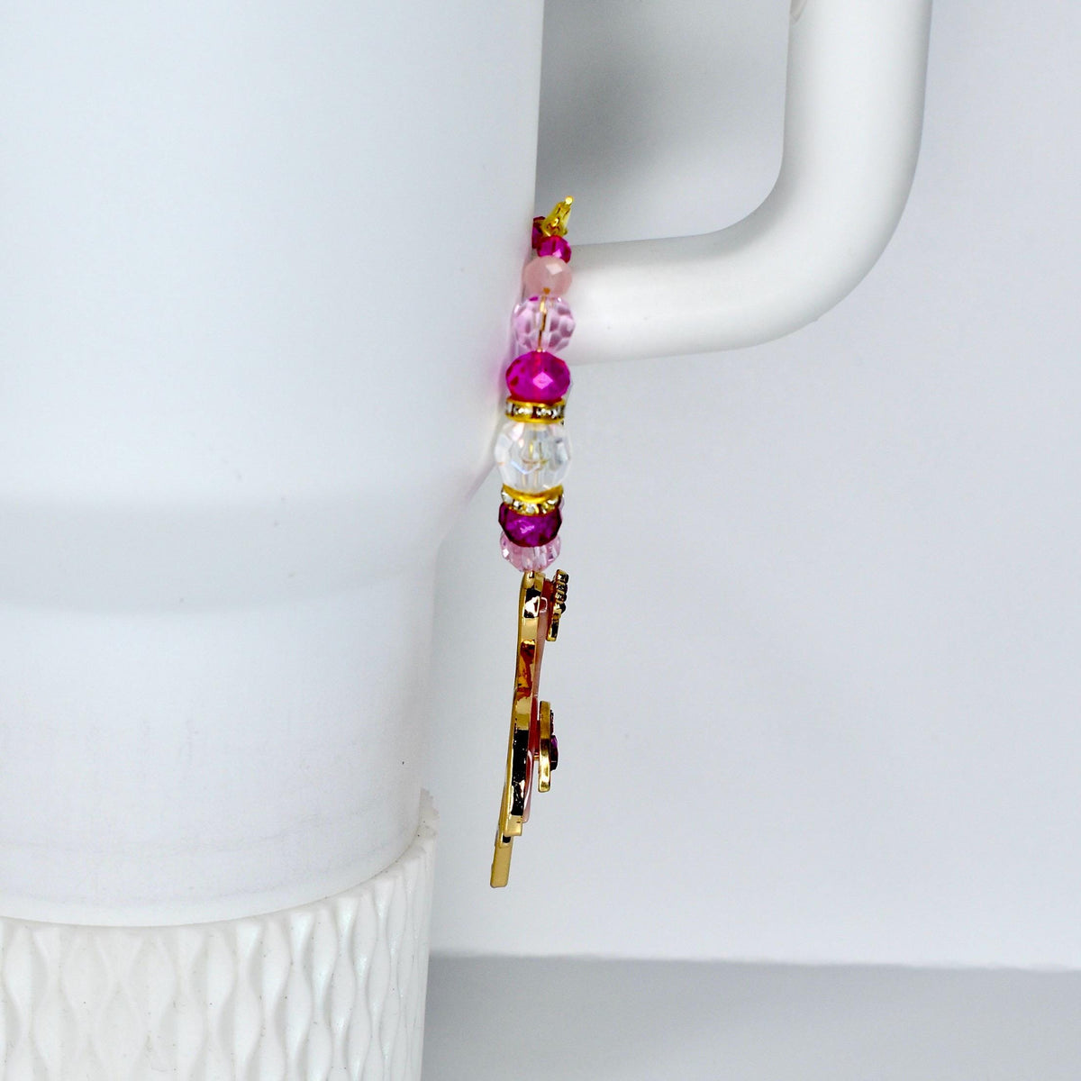 A gold-colored charm with pink and purple gemstones and a beaded strand of pink, white, and purple beads. The charm is hanging on a white mug handle.