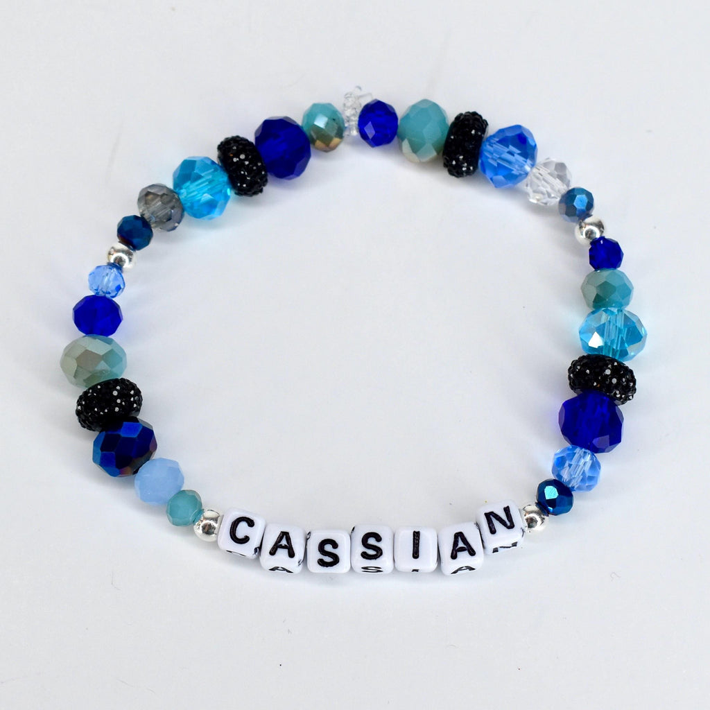 A beaded bracelet with the name "CASSIAN" spelled out in white letter beads. The bracelet features a variety of blue beads, including faceted, round, and crystal beads in shades of light blue, dark blue, and teal. Some beads are black with glitter.