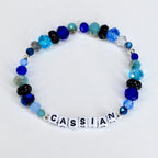 A beaded bracelet with the name "CASSIAN" spelled out in white letter beads. The bracelet features a variety of blue beads, including faceted, round, and crystal beads in shades of light blue, dark blue, and teal. Some beads are black with glitter.