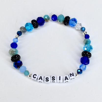 A beaded bracelet with the name "CASSIAN" spelled out in white letter beads. The bracelet features a variety of blue beads, including faceted, round, and crystal beads in shades of light blue, dark blue, and teal. Some beads are black with glitter.