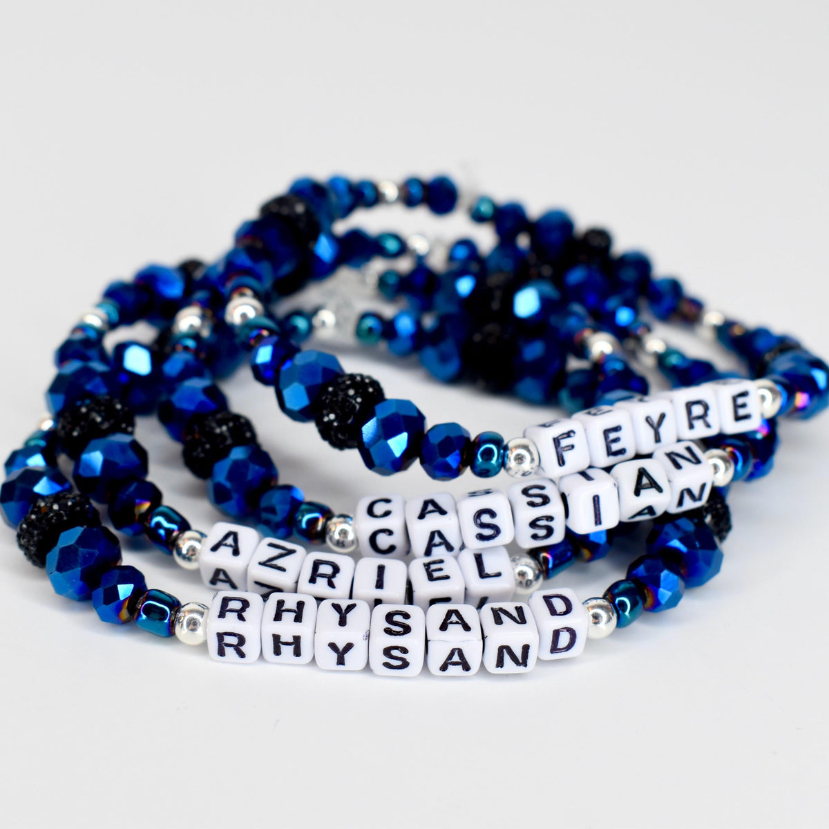 Four beaded bracelets in shades of blue, black, and silver. The bracelets feature faceted beads and small white letter beads spelling out names. The bracelets are stacked together, creating a layered effect.