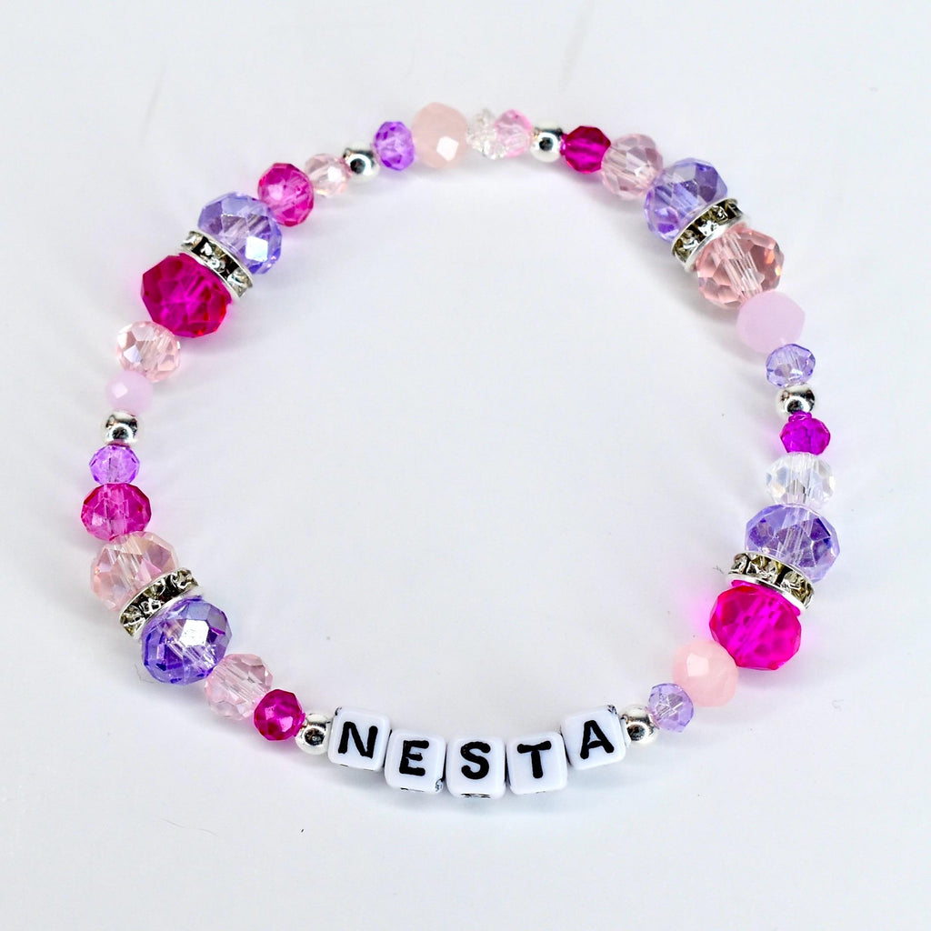 A beaded bracelet with the name "NESTA" spelled out in white letter beads. The bracelet features a variety of pink and purple beads, including faceted and round beads, with silver accents. The beads are strung on a clear elastic cord.