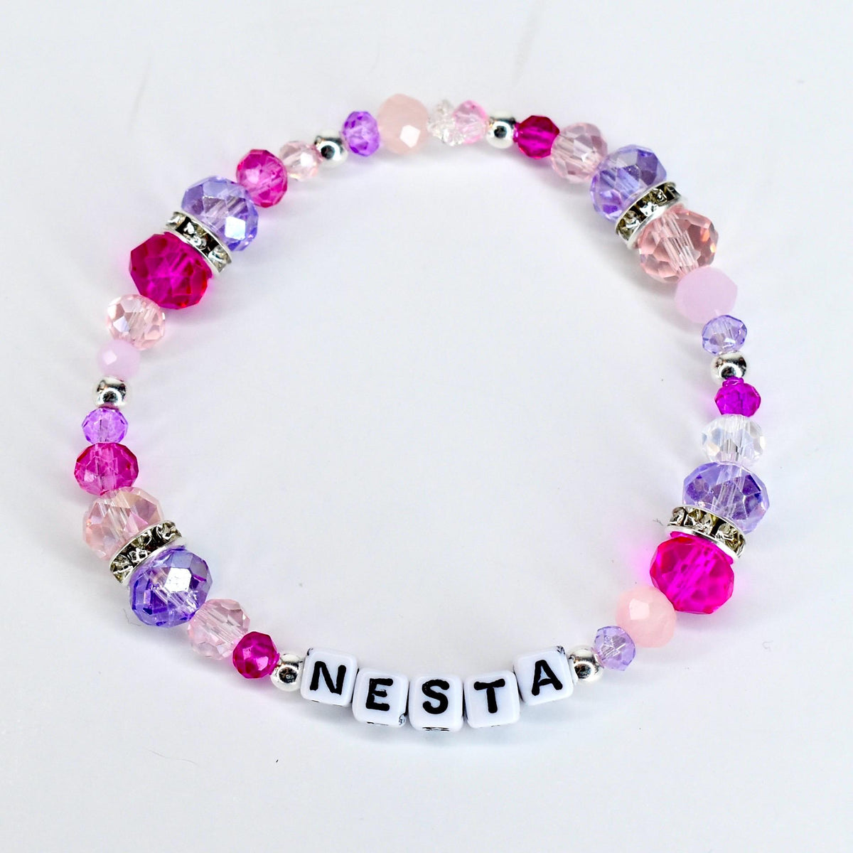 A beaded bracelet with the name "NESTA" spelled out in white letter beads. The bracelet features a variety of pink and purple beads, including faceted and round beads, with silver accents. The beads are strung on a clear elastic cord.