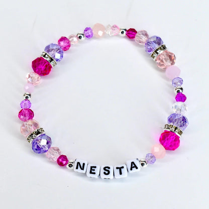 A beaded bracelet with the name "NESTA" spelled out in white letter beads. The bracelet features a variety of pink and purple beads, including faceted and round beads, with silver accents. The beads are strung on a clear elastic cord.