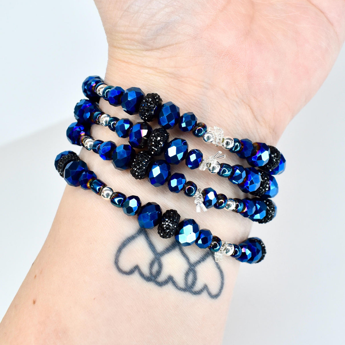 A collection of wrap bracelets featuring faceted blue beads, interspersed with silver-toned beads and black, textured beads. The bracelets are wrapped around a wrist, showcasing the varying textures and colors.