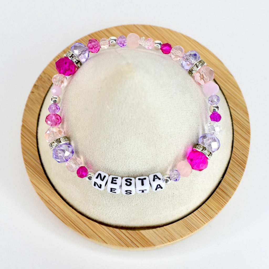 A beaded bracelet with the name "NESTA" spelled out in white letter beads. The bracelet features a variety of pink, purple, and clear beads, including faceted and round shapes. Silver-colored accents are interspersed throughout the design.