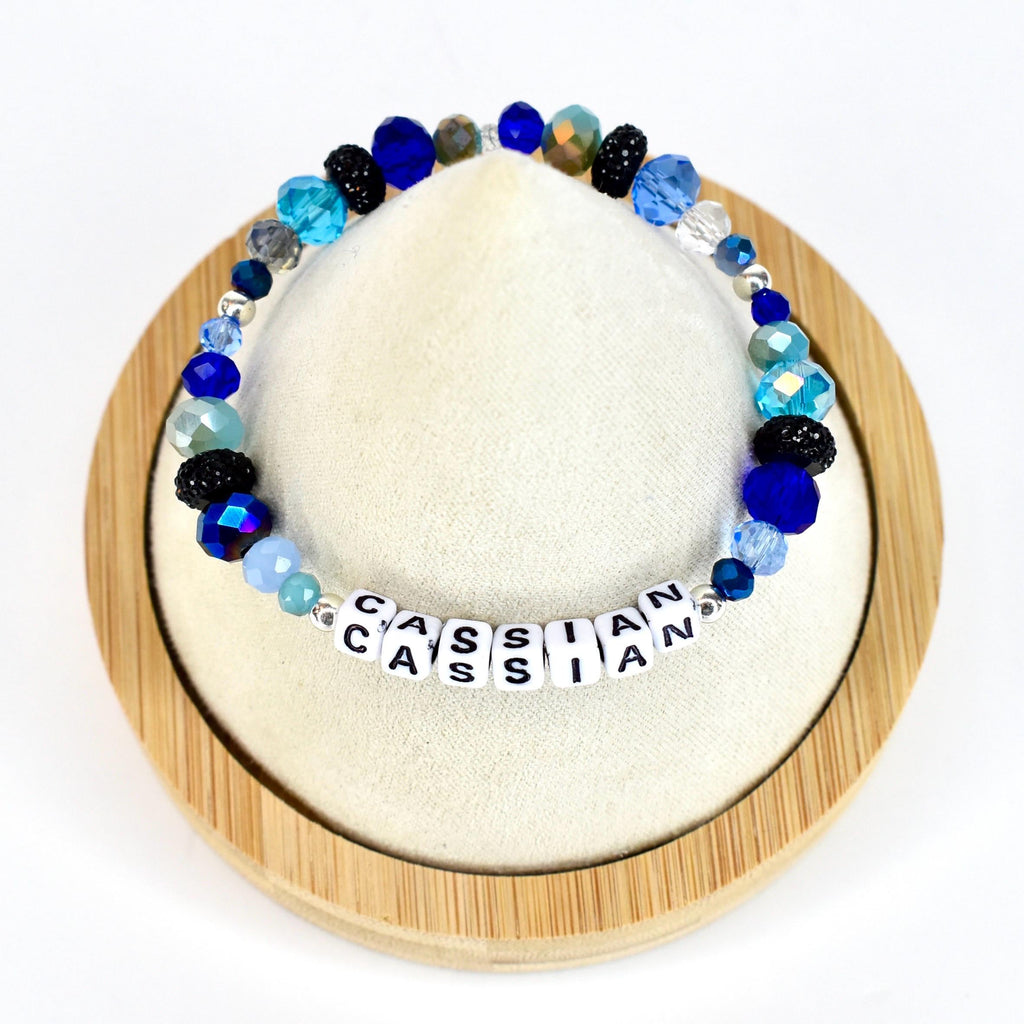 A beaded bracelet with the name "CASSIAN" spelled out in white letter beads. The bracelet features a variety of blue beads, including faceted, round, and crystal-like beads. The bracelet is displayed on a white surface.