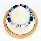 A beaded bracelet with the name "CASSIAN" spelled out in white letter beads. The bracelet features a variety of blue beads, including faceted, round, and crystal-like beads. The bracelet is displayed on a white surface.